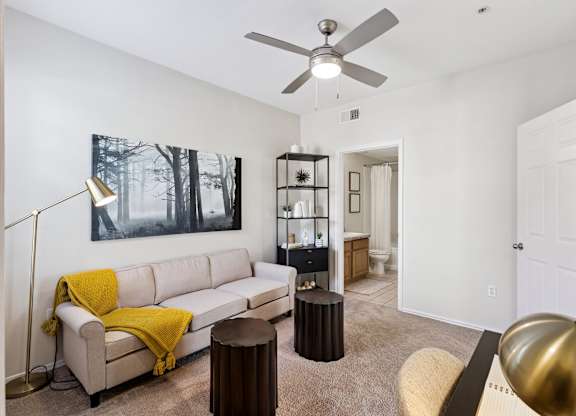 A home office here at Quail Landing with soft carpet flooring, a beige sofa accented by a mustard throw, black shelving, small round stools, a ceiling fan, and warm natural light creating a flexible, inviting workspace.