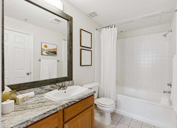 A bathroom here at Quail Landing featuring a granite vanity with wood cabinetry, large framed mirror, white toilet, and a tub with white tile surround and shower curtain, styled with simple decor and soft lighting.