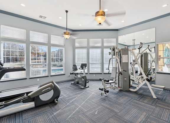 A fitness center here at Quail Landing with modern cardio and strength equipment, blue patterned flooring, ceiling fans, and large windows that bring in bright natural light.
