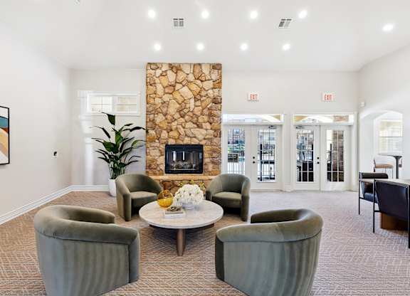 A community lounge here at Quail Landing with four soft green lounge chairs around a round coffee table, a tall stone fireplace, light carpet flooring, large windows, and bright recessed ceiling lighting.