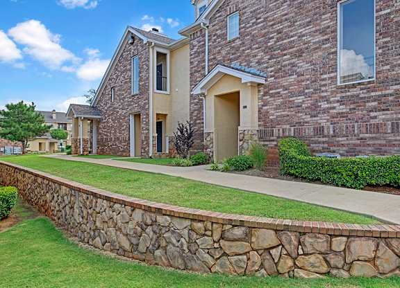 A community exterior walkway here at Quail Landing with brick apartment buildings, stone retaining wall, green landscaping, covered entryways, and a clean concrete path under a bright blue sky.