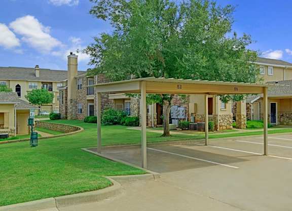 A covered parking area here at Quail Landing with carport spaces, green lawns, mature trees, and surrounding apartment buildings, offering a clean, convenient, and well-maintained community setting.
