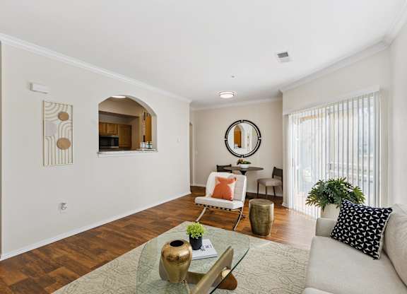 A living room here at Quail Landing with warm wood floors, a beige sofa with black patterned pillows, a glass coffee table on a light rug, a small dining nook, leafy green plants, and bright sliding glass doors.