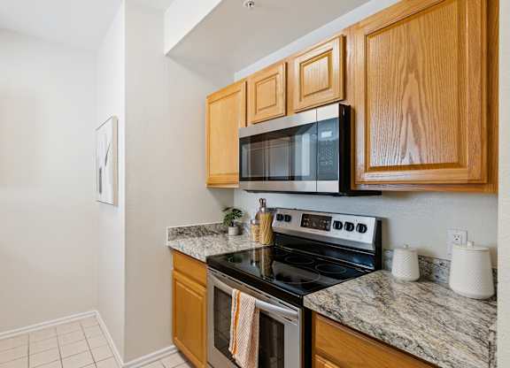 A kitchen here at Quail Landing with warm wood cabinets, granite-style countertops, stainless steel stove and microwave, light tile flooring, and a clean, well-lit layout.