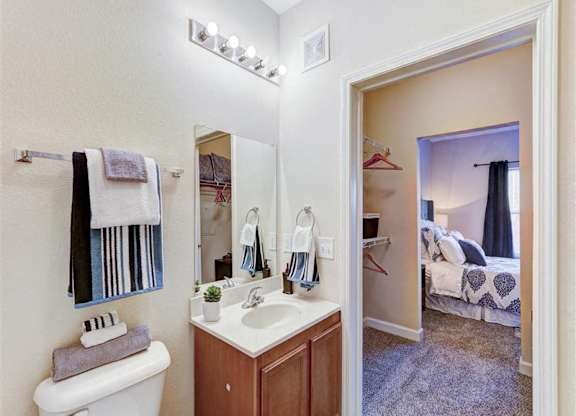 A bathroom with a toilet, sink, mirror, and a bedroom in the background.