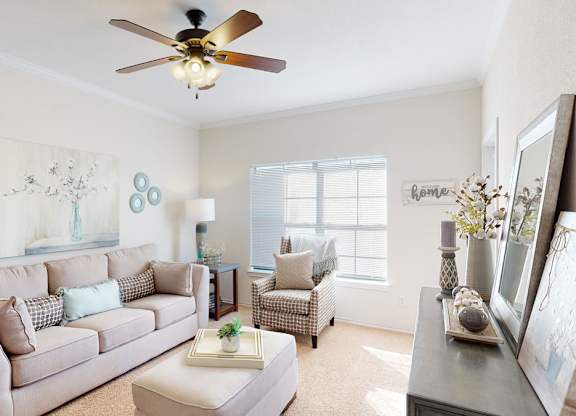 A bright living room here at Stoneleigh on May with beige carpet, a soft neutral sofa, a patterned accent chair, and a ceiling fan. Sunlight filters through wide windows, creating a warm, inviting, and comfortable atmosphere.
