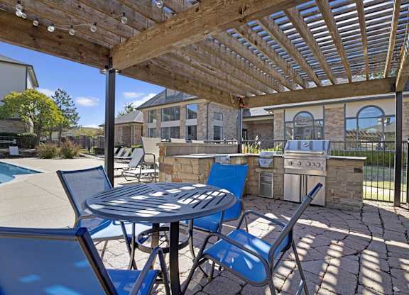 A shaded outdoor grilling area here at Stoneleigh on May with a pergola, built-in grills, and stone countertops. Patio seating nearby and views of the pool create a relaxed, social space for outdoor dining and gatherings.