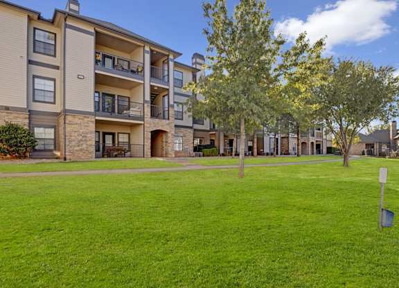 A landscaped community exterior here at Stoneleigh on May with manicured green lawns, mature trees, and multi-story apartment buildings with balconies. Stone accents and walking paths create a welcoming, park-like residential setting.