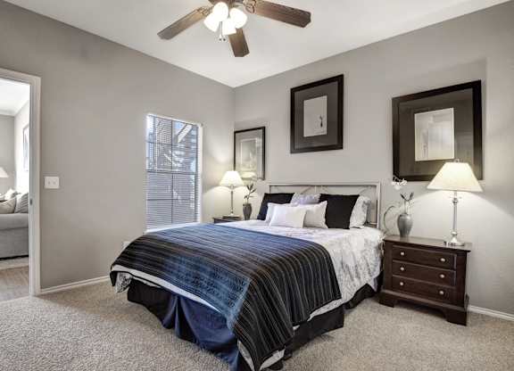 A comfortable bedroom here at Stoneleigh on May with beige carpet, soft gray walls, and a ceiling fan with light. A large bed with neutral bedding is flanked by matching nightstands, while a window with blinds brings in natural light.