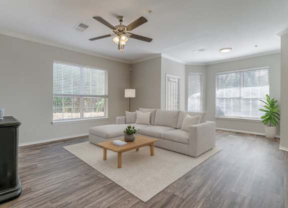 A bright living room here at Sycamore Farms with wood-style flooring, a neutral sectional sofa, and a ceiling fan with light. Large windows with blinds bring in natural light, creating an open, calm, and inviting atmosphere.