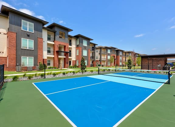 A tennis court here at The Encore with bright blue and green playing surfaces, modern netting, and landscaped surroundings. The open-air court sits alongside contemporary apartment buildings under a clear blue sky.