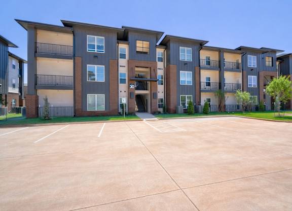 A apartment building exterior here at The Encore with modern three-story architecture, gray and brick accents, private balconies, and landscaped grounds under a clear blue sky.