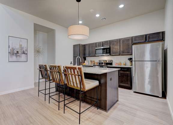 A kitchen here at The Encore with a spacious island, dark wood cabinetry, and stainless steel appliances. Light flooring, a pendant light, and wood-tone bar stools create a warm, modern gathering space.
