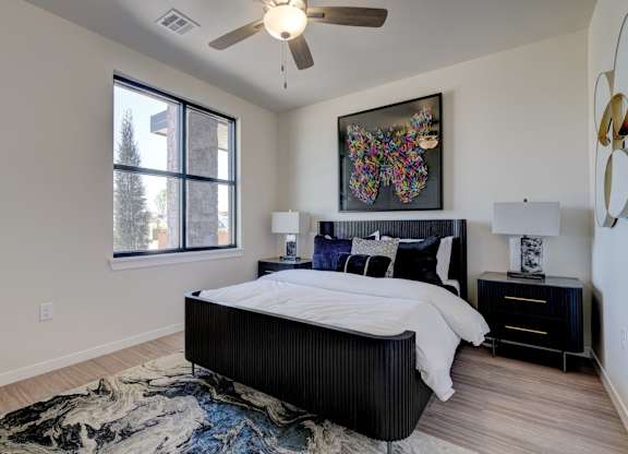 A bedroom here at The Encore with a black bed frame, crisp white bedding, and colorful wall art above the headboard. Light flooring, matching nightstands with lamps, a ceiling fan, and a large window create a bright, modern feel.