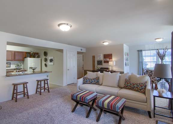 A living room with a couch, a striped ottoman, and a kitchen area in the background.
