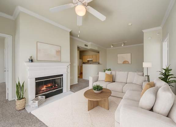 A living room here at Woodland Park with a light beige sofa set, cream rug, and round wood coffee table, centered around a white fireplace, with soft green walls, crown molding, and a white ceiling fan overhead.