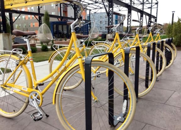 A row of bright yellow bicycles is lined up neatly at a bike rack outdoors, with an industrial building in the background, outside Stitchweld Apartments.