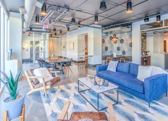 Modern lounge with a stylish blue sofa, geometric rug, and wooden chairs. Exposed ceiling pipes and patterned walls create a chic, inviting atmosphere at Stitchweld Apartments.
