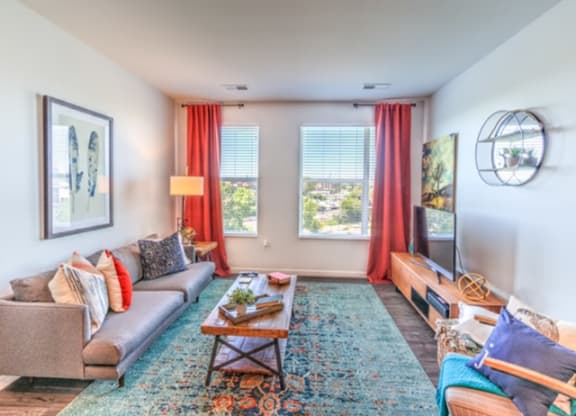 A bright, cozy living room with a gray couch, colorful cushions, wooden coffee table, large rug, vibrant red curtains, and a flat-screen TV at Stitchweld Apartments.