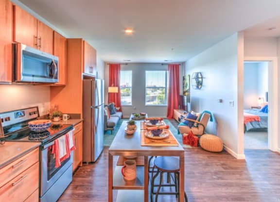 Modern kitchen with wooden cabinets, stainless steel appliances, and a central island set for dining. The cozy living area has vibrant red curtains and soft lighting at Stitchweld Apartments.