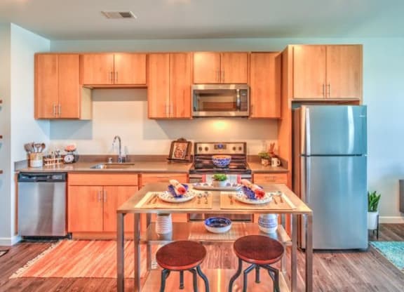 A cozy kitchen with wooden cabinets, a stainless steel fridge, microwave, and a small dining table set for two at Stitchweld Apartments.