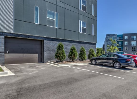 Parking lot with gray building on the left, featuring small trees lined along the side. A parked black car is visible under a clear blue sky at Stitchweld Apartments.