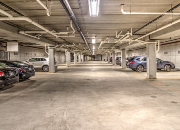Dimly lit underground parking garage with concrete pillars and cars parked on both sides. The space appears orderly and empty, conveying a quiet atmosphere at Stitchweld Apartments.