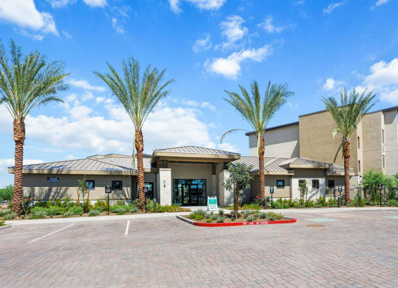 Modern Reflect at Dobson Ranch Apartment building with beige exteriors, palm trees, and clear blue sky. The foreground shows an empty brick parking lot and a "Now Open" sign.