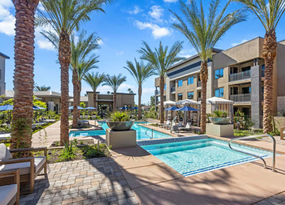 Outdoor area here at Reflect at Dobson Ranch Apartments with palm trees and a sparkling pool under a clear blue sky. Lounge chairs and umbrellas create a relaxing, tropical atmosphere.