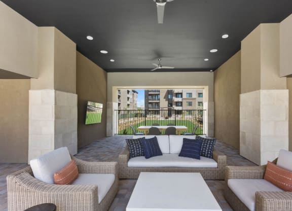 Outdoor patio area here at Reflect at Dobson Ranch Apartments with wicker furniture, colorful cushions, and a central white table. Background shows modern buildings and a grassy courtyard.
