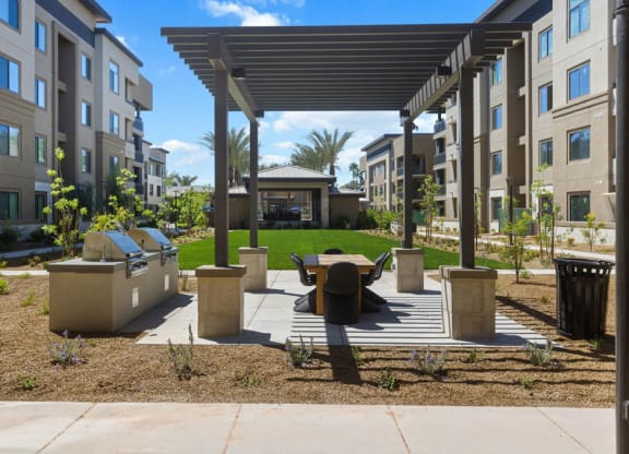 Outdoor Reflect at Dobson Ranch Apartment courtyard with a modern pergola, picnic table, and barbecue grills. Buildings flank lush lawns, conveying a welcoming, communal vibe.