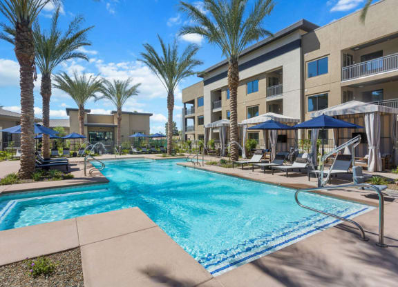 Luxurious pool area here at Reflect at Dobson Ranch Apartments with palm trees, cabanas, and lounge chairs under blue umbrellas. Modern apartment building in the background under a sunny sky.