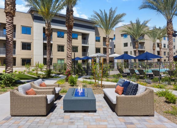 Outdoor seating area here at Reflect at Dobson Ranch Apartments with wicker sofas and colorful cushions surrounds a modern fire pit. Tall palm trees and a pool with umbrellas are in the background.