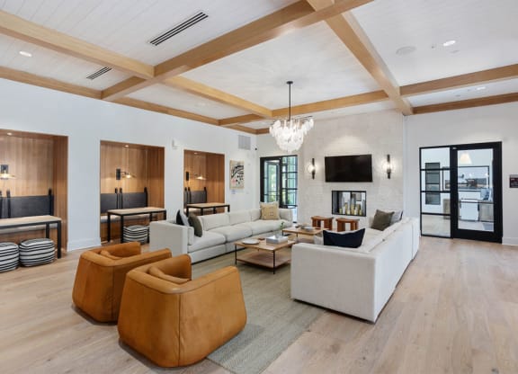 Modern lobby here at Reflect at Dobson Ranch Apartments with white walls, wood ceiling beams, and light wood flooring. White sofas, brown chairs, and a central coffee table create a cozy, elegant space. A TV and fireplace are set against a textured wall, while booths with striped cushions are nearby. Large windows allow natural light.