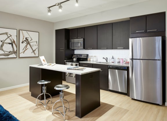 Modern kitchen here at 4Marq Apartment Homes with dark cabinets, stainless steel appliances, and a sleek island with two silver stools. Abstract art and hardwood floors add style.