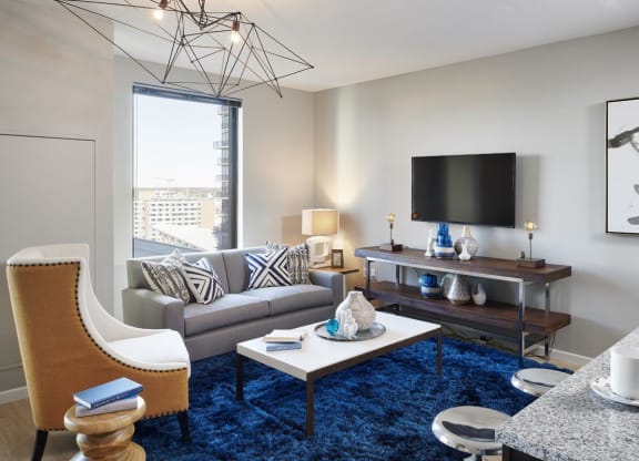 Modern living room here at 4Marq Apartments with a gray sofa adorned with geometric pillows, a blue shag rug, and a sleek coffee table. Wall-mounted TV, abstract art, and decorative vases add elegance.