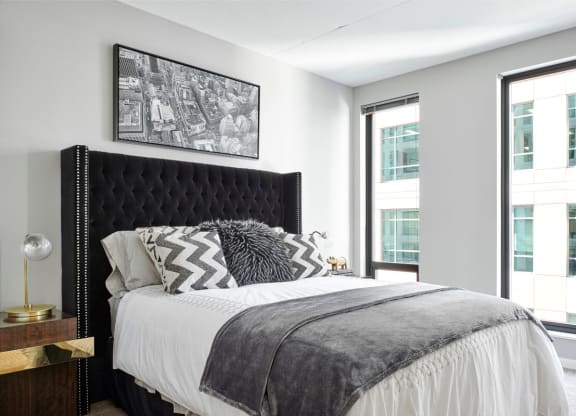 Modern bedroom here at here at 4Marq Apartments with a black tufted headboard, monochrome zigzag and furry pillows, gray throw blanket, bedside table, lamp, and cityscape photo. Bright and cozy ambiance.