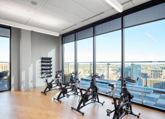 Modern gym room here at 4Marq Apartments with three stationary bikes on wooden floor. Large windows offer a panoramic city view. Bright, clean, and motivational.