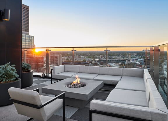 Rooftop patio here at 4Marq Apartments at sunset, featuring a modern gray sectional sofa surrounding a square fire pit. Glass railings provide a cityscape view, creating a serene ambiance.