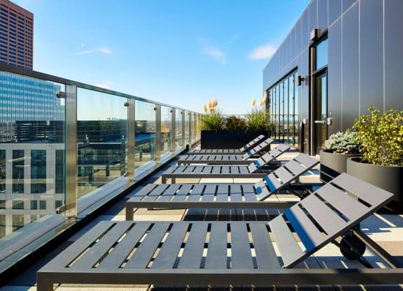 Rooftop terrace here at 4Marq Apartments with sleek lounge chairs lined up under a clear blue sky. Glass railing offers city views, and planters with greenery add elegance.