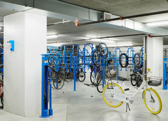Underground bike parking here at 4Marq Apartments with blue racks holds various bicycles. A single white bike with yellow tires and a front basket stands out in the foreground.