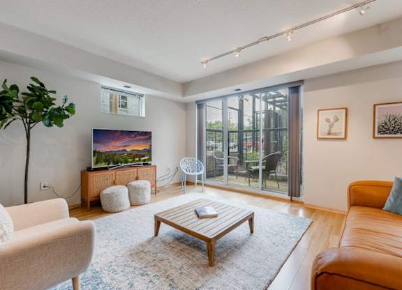 Modern living room here at Blue and Lime Apartments with a comfy brown leather sofa and white armchair. Large TV on a wooden stand, potted plant, and sliding glass doors to a patio.