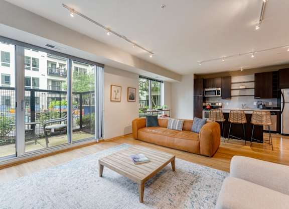 Modern living room here at Blue and Lime Apartments with a tan leather sofa, wooden coffee table, open kitchen, and large windows with a view of greenery. Bright and cozy atmosphere.