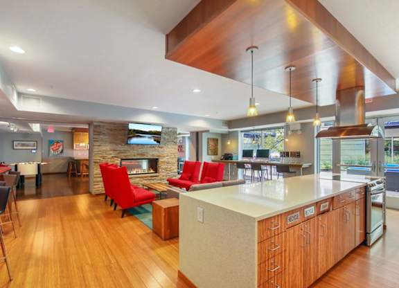 Modern community lounge here at Blue and Lime Apartments with bamboo floors, stone fireplace, and vibrant red chairs. A kitchen island with pendant lights and an outdoor view provides a cozy feel.