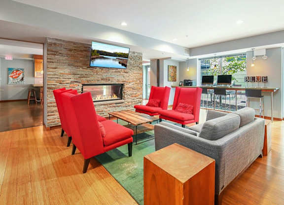 Modern lounge here at Blue and Lime Apartments with vibrant red chairs and a gray sofa around a wooden coffee table. A stone fireplace and large windows add warmth and light.