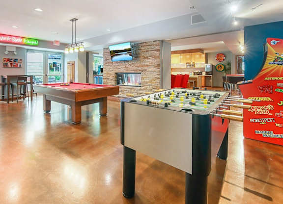 Game room here at Blue and Lime Apartments with a red pool table and foosball table on a polished floor. A stone fireplace with a TV is in the background. Bright, casual atmosphere.