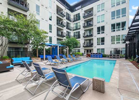 Modern courtyard here at Blue and Lime Apartments with a rectangular pool surrounded by blue lounge chairs and seating. Tall building with balconies and greenery. Bright and inviting.