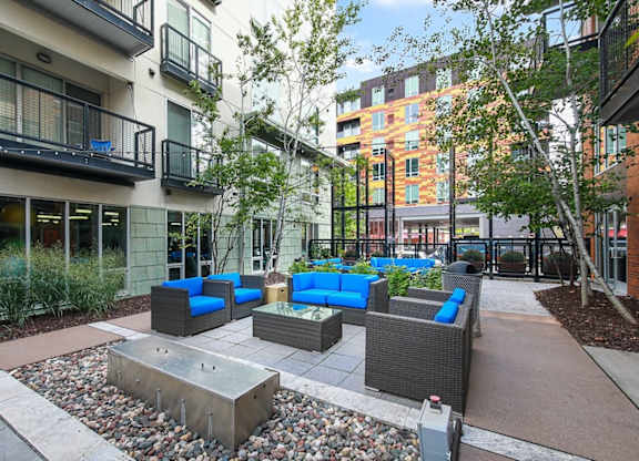 Modern outdoor courtyard here at Blue and Lime Apartments with wicker furniture featuring bright blue cushions. Surrounded by apartment buildings, trees, and a stone path, creating a vibrant, inviting atmosphere.