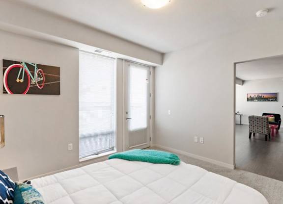 Modern bedroom here at Blue and Lime Apartments with a crisp white bed and teal accents, featuring a bicycle wall art. A door opens to a bright living room with colorful chairs.