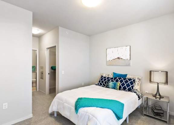 A modern bedroom here at Blue and Lime Apartments with a neatly made bed featuring a white comforter and teal accents. A lamp sits on a bedside table, and a bathroom is seen in the background.